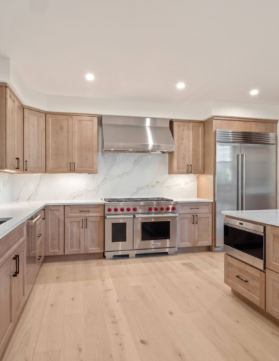 A modern kitchen featuring wooden cabinetry, a large island, stainless steel appliances, and marble countertops, illuminated with bright overhead lighting.