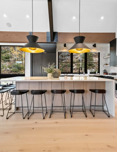 A modern kitchen with an island, bar stools, and stylish pendant lights, featuring large windows and a blend of black and white cabinetry.