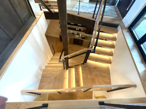 A modern staircase featuring illuminated steps, wooden finishes, and a sleek design, leading down from an upper level to a lower level.