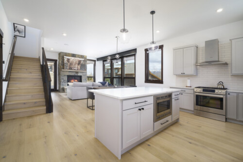 A modern kitchen with a central island, stainless steel appliances, large windows, and wooden flooring, featuring a staircase in the background.