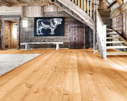 A rustic wooden interior features a large staircase, a cow illustration on the wall, and a warm wooden floor.