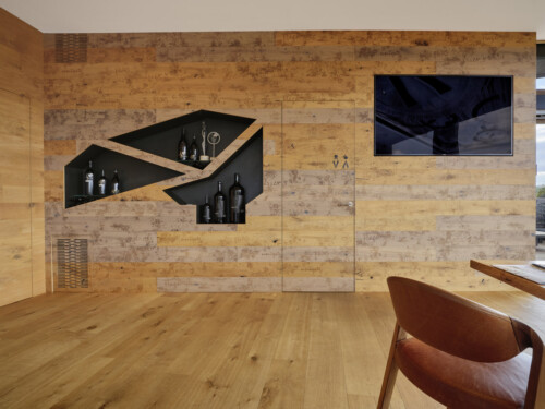A modern interior with wooden walls, featuring a unique angular shelf displaying glass bottles and a television mounted above.