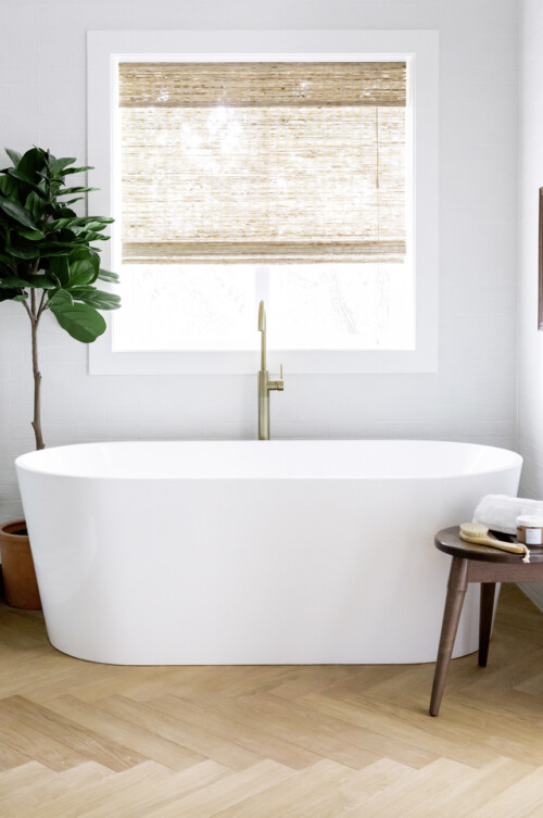 A modern bathroom featuring a white freestanding bathtub, a wooden floor, and a potted plant beside a window with woven shades.