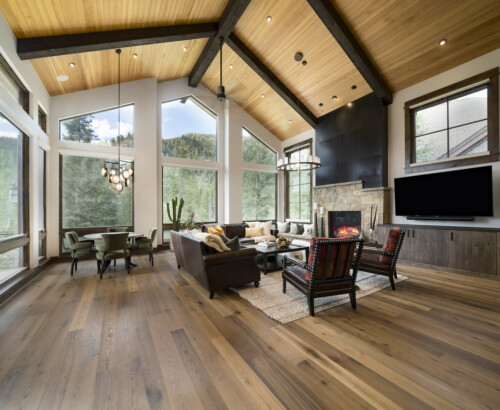 A spacious, modern living room with large windows, wooden beams, and elegant furnishings, featuring a cozy fireplace and scenic outdoor views.