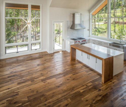 A modern kitchen with large windows, wooden flooring, and minimalist design features, illuminated by natural light.