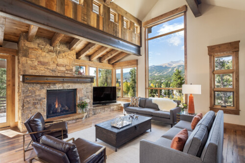 A spacious, modern living room with large windows overlooking mountains, featuring a stone fireplace, comfortable seating, and warm wood accents.
