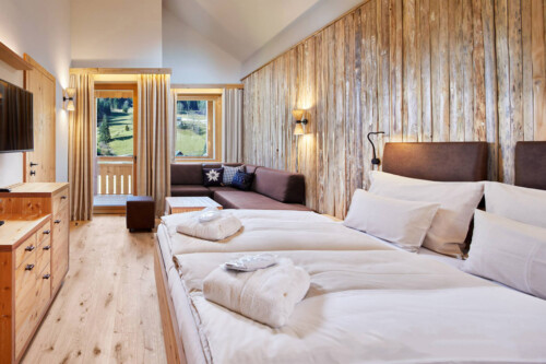 A cozy, modern bedroom featuring wooden walls, two beds, a seating area, and large windows allowing natural light.