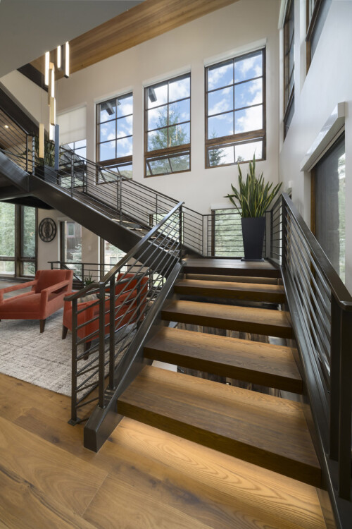 A stylish interior featuring a wooden staircase, modern railings, large windows, and a contemporary living space with red seating and green plants.