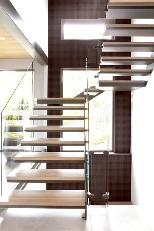 A modern staircase with wooden steps and glass railings, illuminated by natural light from large windows.